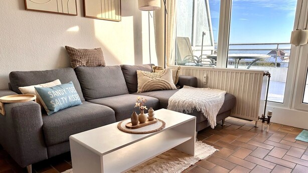 A modern living room with a grey sofa, a white coffee table, and a view of the sea through large windows.