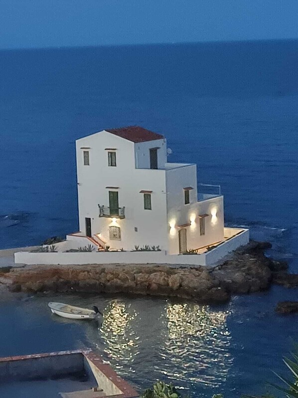 A modern white building with balconies, situated on a rocky outcrop by the sea.
