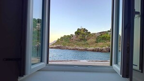 A window with a view of a coastal landscape, including a body of water and a rocky hillside with vegetation. - Casetta Bianca Luxury Santa Flavia (Santa Flavia)