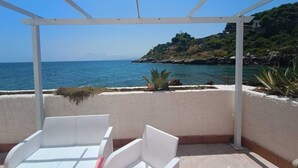 A covered outdoor seating area with white wicker furniture, overlooking a coastal landscape with a clear view of the sea and a rocky hillside. - Casetta Bianca Luxury Santa Flavia (Santa Flavia)