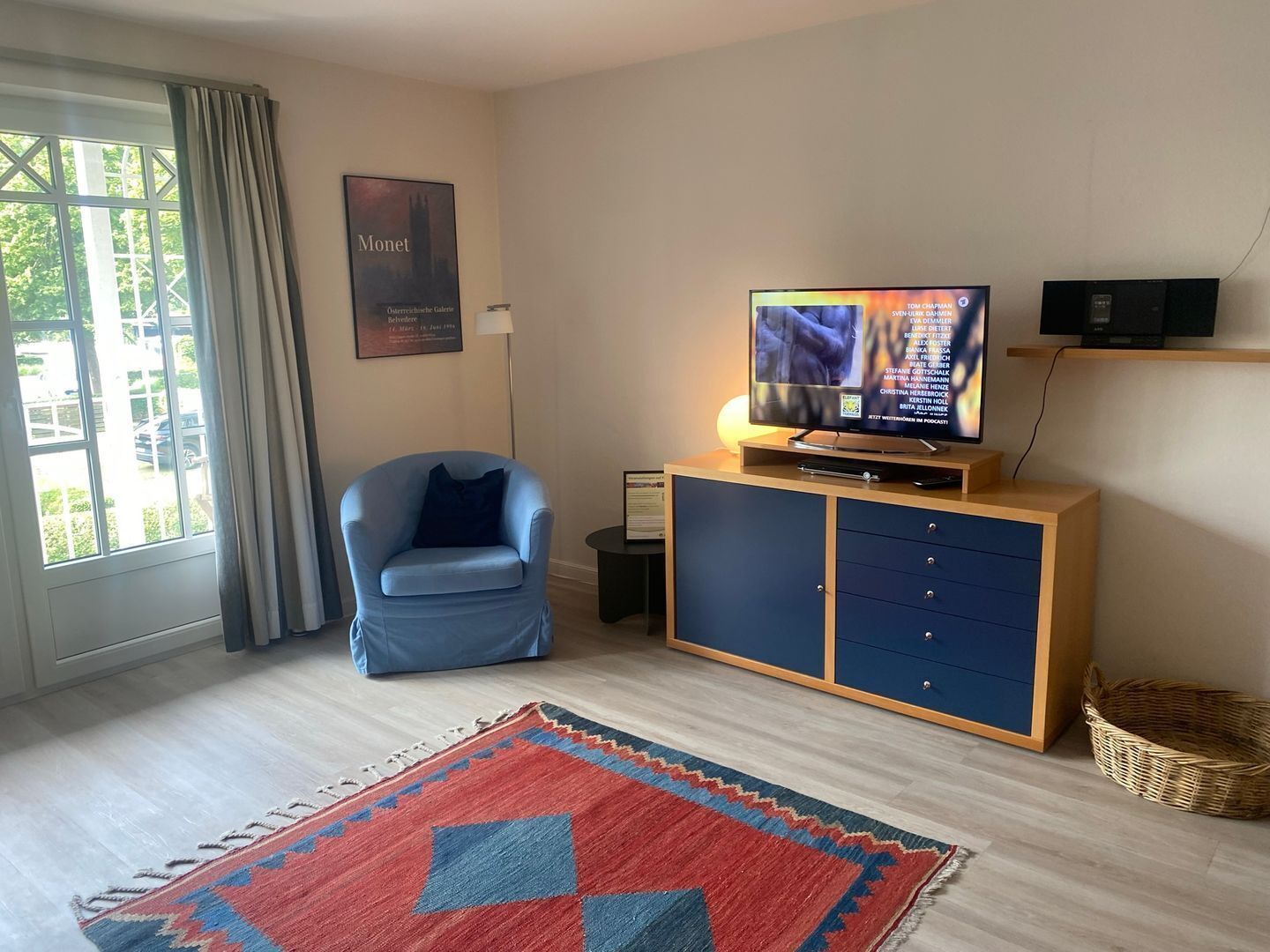 Una sala de estar moderna con un sillón azul, un televisor sobre un soporte de madera, un tapete estampado y un cuadro enmarcado en la pared.