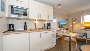 A modern kitchen with white cabinets, a Bosch microwave, and a coffee maker. There is a dining area with a table and chairs, and a view of the sea through the window. - Gusy 4 U - 1-room vacation apartment Gusy 4 U in the heart of Westerland, 100 m from the (Sylt)
