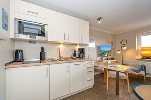 A modern kitchen with white cabinets, a Bosch microwave, and a coffee maker. There is a dining area with a table and chairs, and a view of the sea through the window.