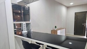 Een moderne keuken met een zwart aanrecht, witte kasten en een ingebouwde oven. Twee zwarte barkrukken en een klein houten bijzettafeltje met een potplant.