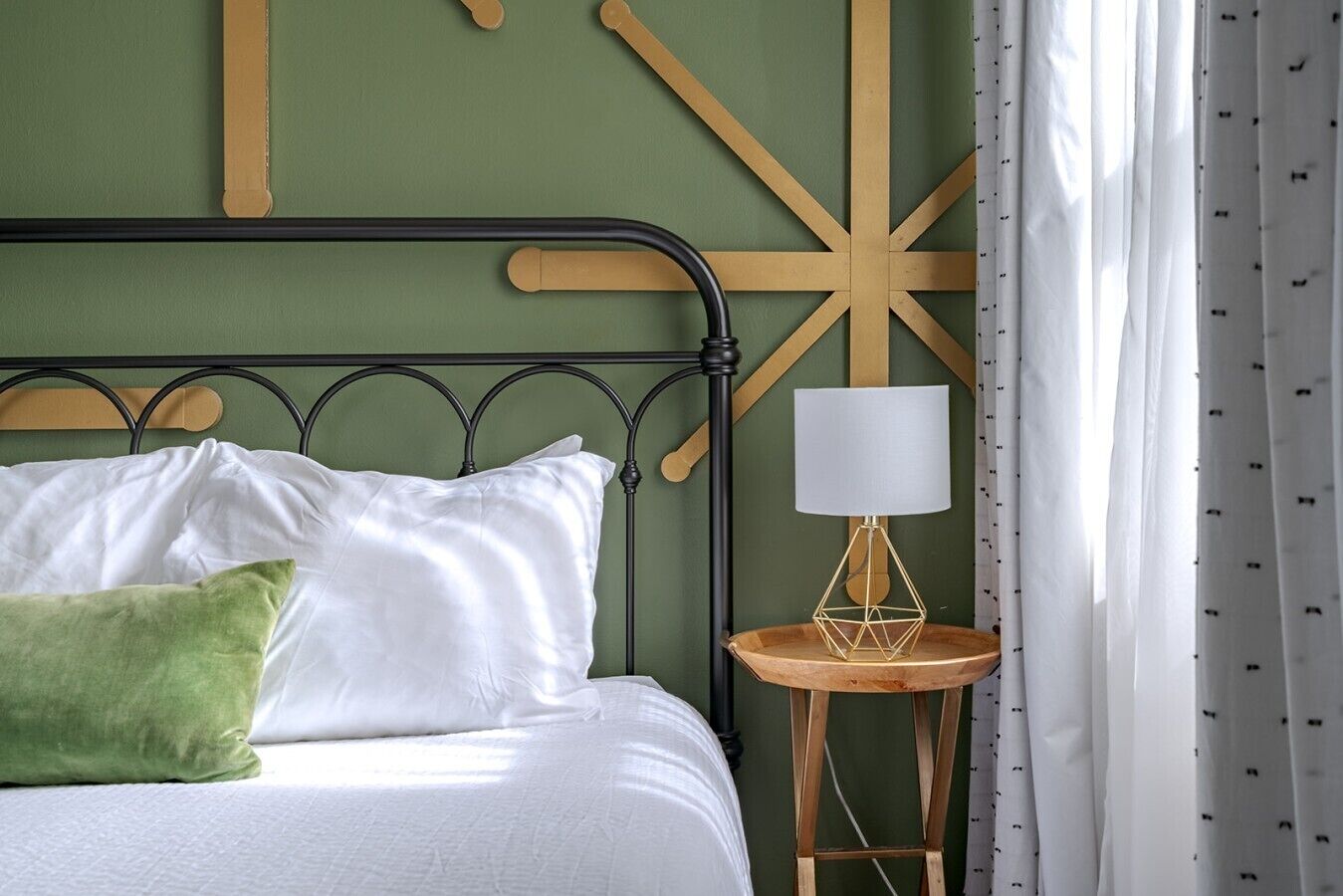 Ein Bett mit weißer Bettwäsche und einem grünen Kopfkissen, eine Nachttischlampe und eine Wand mit geometrischen Mustern.