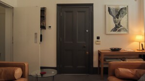 A living room with a dark wooden door, a glass table, two leather sofas, a small round table, and a framed artwork on the wall.