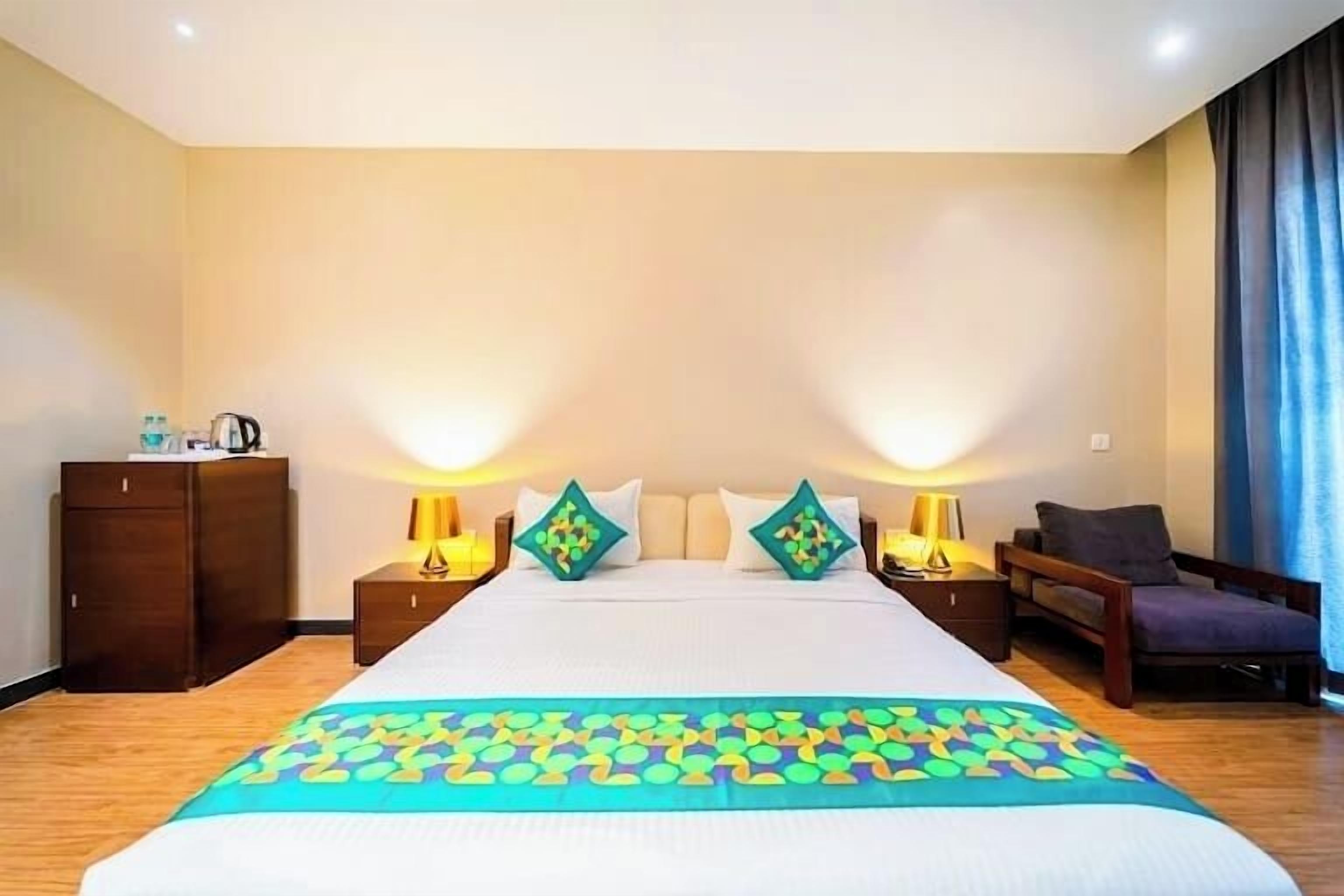 A hotel room with a large bed, two bedside tables with lamps, a wooden wardrobe, a chair, and a blue curtain.