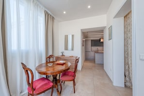 A dining area with a round wooden table, red cushioned chairs, a kitchen in the background, and a large mirror on the wall.