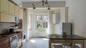 A kitchen with a stove, microwave, and washing machine. There is a window with curtains, a table with chairs, and a refrigerator. - Giardino Degli Aranci With Terrace by Wonderful Italy (Taormina)