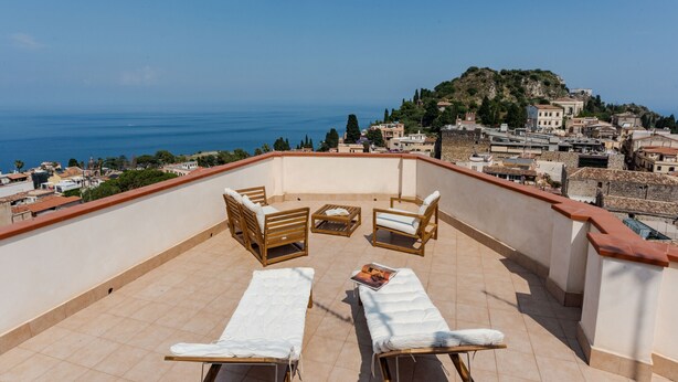 A rooftop terrace with two sun loungers, a small table, and a magazine. The terrace overlooks a coastal town with buildings and a hill in the distance. - Giardino Degli Aranci With Terrace by Wonderful Italy (Taormina)
