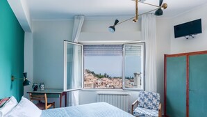 A bedroom with a bed, a chair, a desk, a radiator, a window with curtains, a TV, and a wardrobe. - Giardino Degli Aranci With Terrace by Wonderful Italy (Taormina)