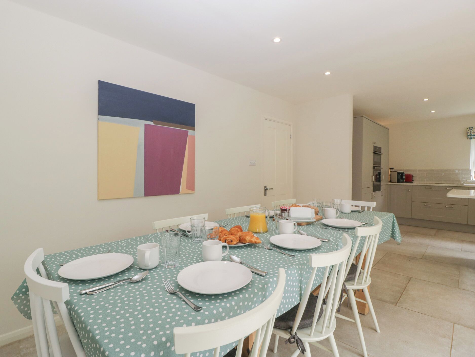 Un comedor con una mesa dispuesta para cuatro, sillas blancas y un cuadro abstracto de colores en la pared.