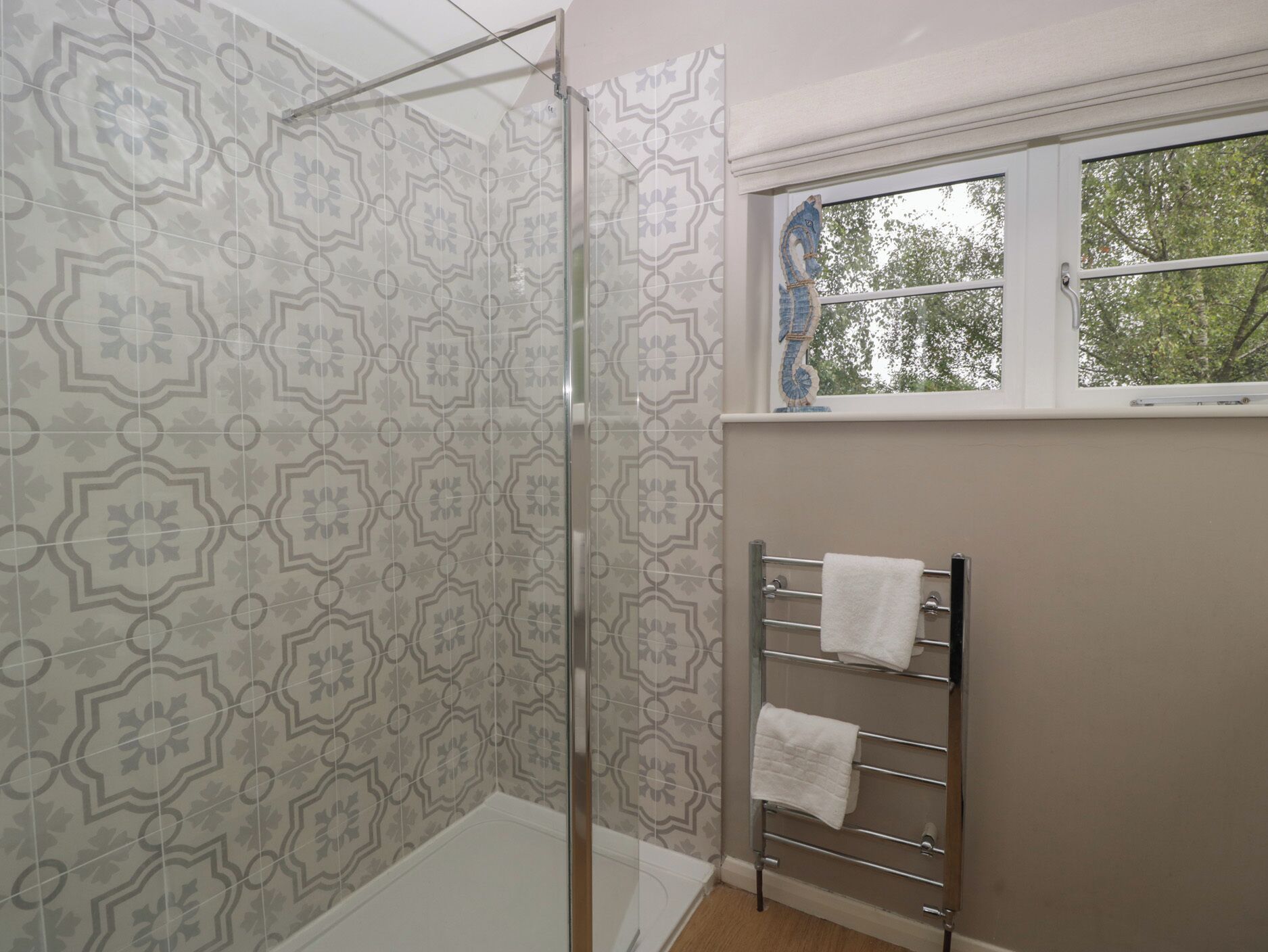 Un baño con baldosas estampadas, cabina de ducha con mampara de vidrio, toallero con dos toallas y ventana con vistas a árboles.
