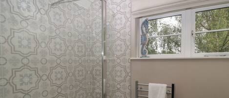 Un baño con baldosas estampadas, cabina de ducha con mampara de vidrio, toallero con dos toallas y ventana con vistas a árboles.