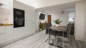 Eine moderne Küche mit eingebautem Ofen und Mikrowelle, ein Essbereich mit Glastisch und grauen Polsterstühlen sowie ein an der Wand befestigter Fernseher.