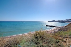 A coastal beach with a pier, clear blue water, and a hilly landscape in the background.