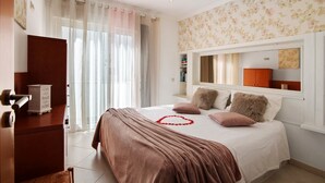 A bedroom with a bed, a nightstand, a dresser, a window with curtains, and a wall with floral wallpaper. - Apartment 'Vista Mar' with Sea View, Wi-Fi and Air Conditioning (Albufeira)