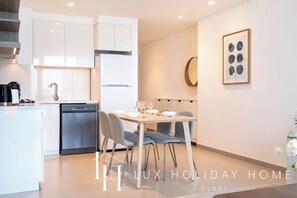 A modern kitchen with white cabinetry, a dishwasher, and a dining area with a table and chairs. - LUX The Downtown Zabeel View Suite (Dubai)