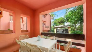 A room with a white table and wicker chairs, a window with a view of greenery, and a balcony with a wooden railing. - Apartment 'Don Carlos Marbella Beach' with Shared Pool, Wi-Fi and Air Conditioning (Marbella)