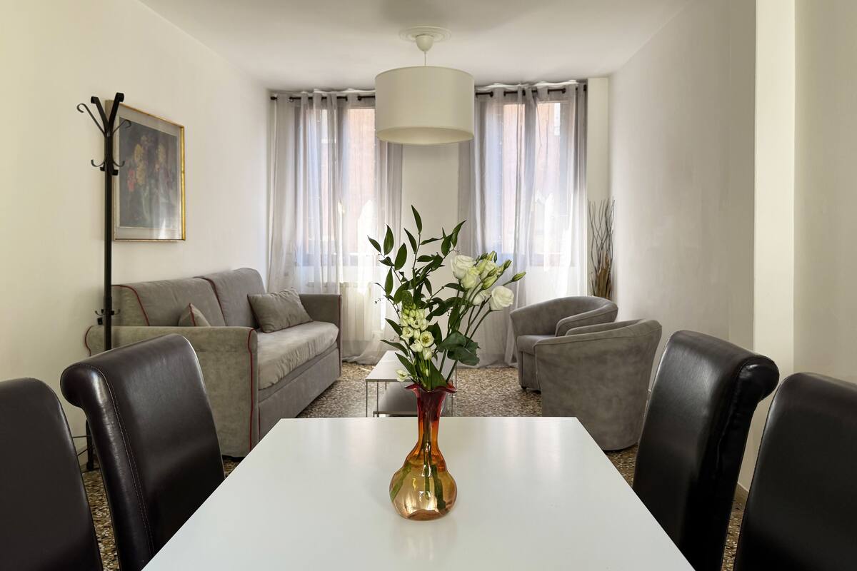 A modern living room with a white table, black leather chairs, a grey sofa, and a vase with white flowers.
