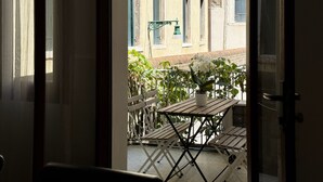 Uma porta aberta para uma varanda com mesa e cadeiras, um vaso de planta e vista de edifícios ao fundo.