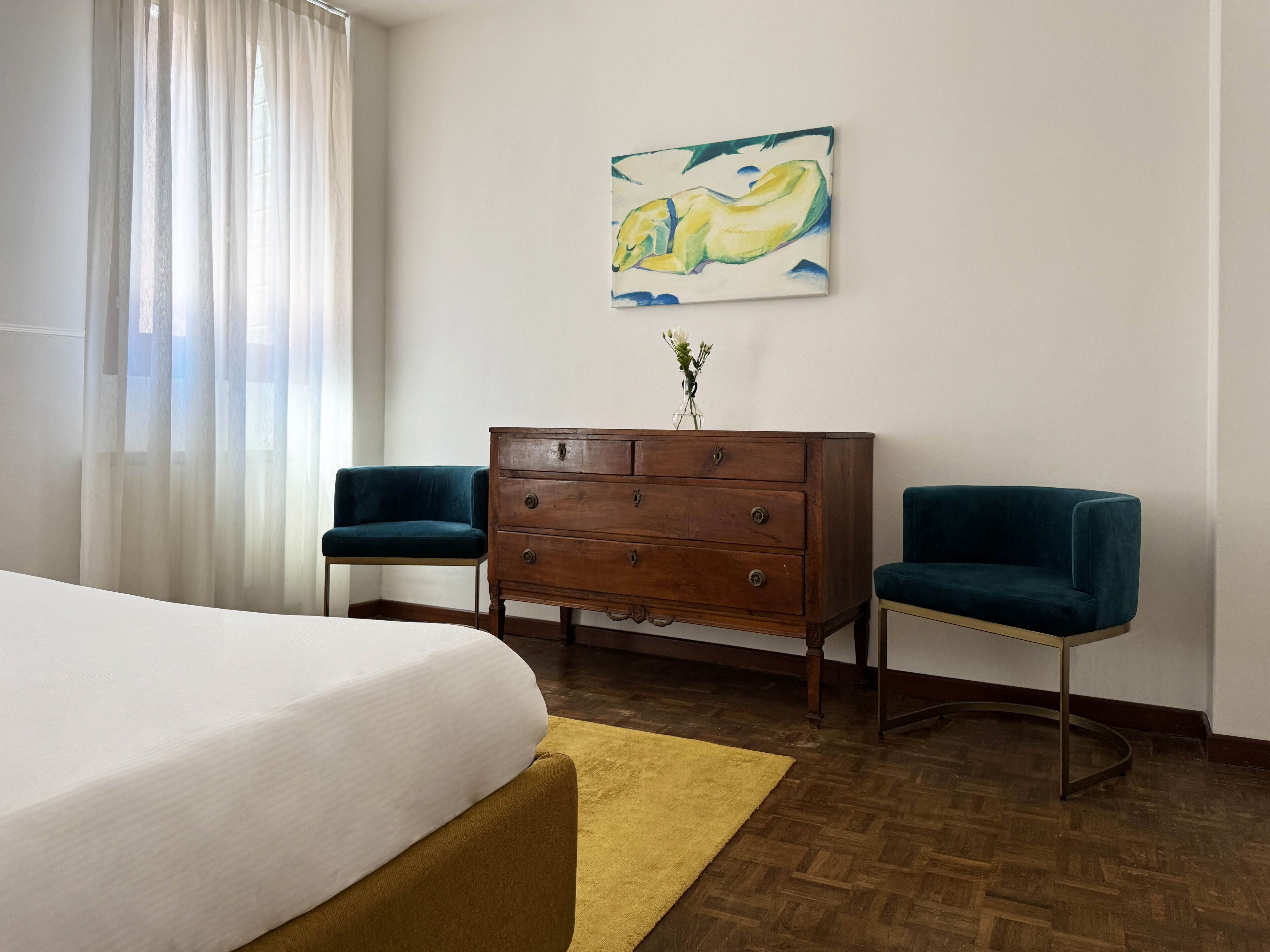Een hotelkamer met een bed, een houten dressoir, twee fauteuils, een schilderij aan de muur en een raam met dunne gordijnen.