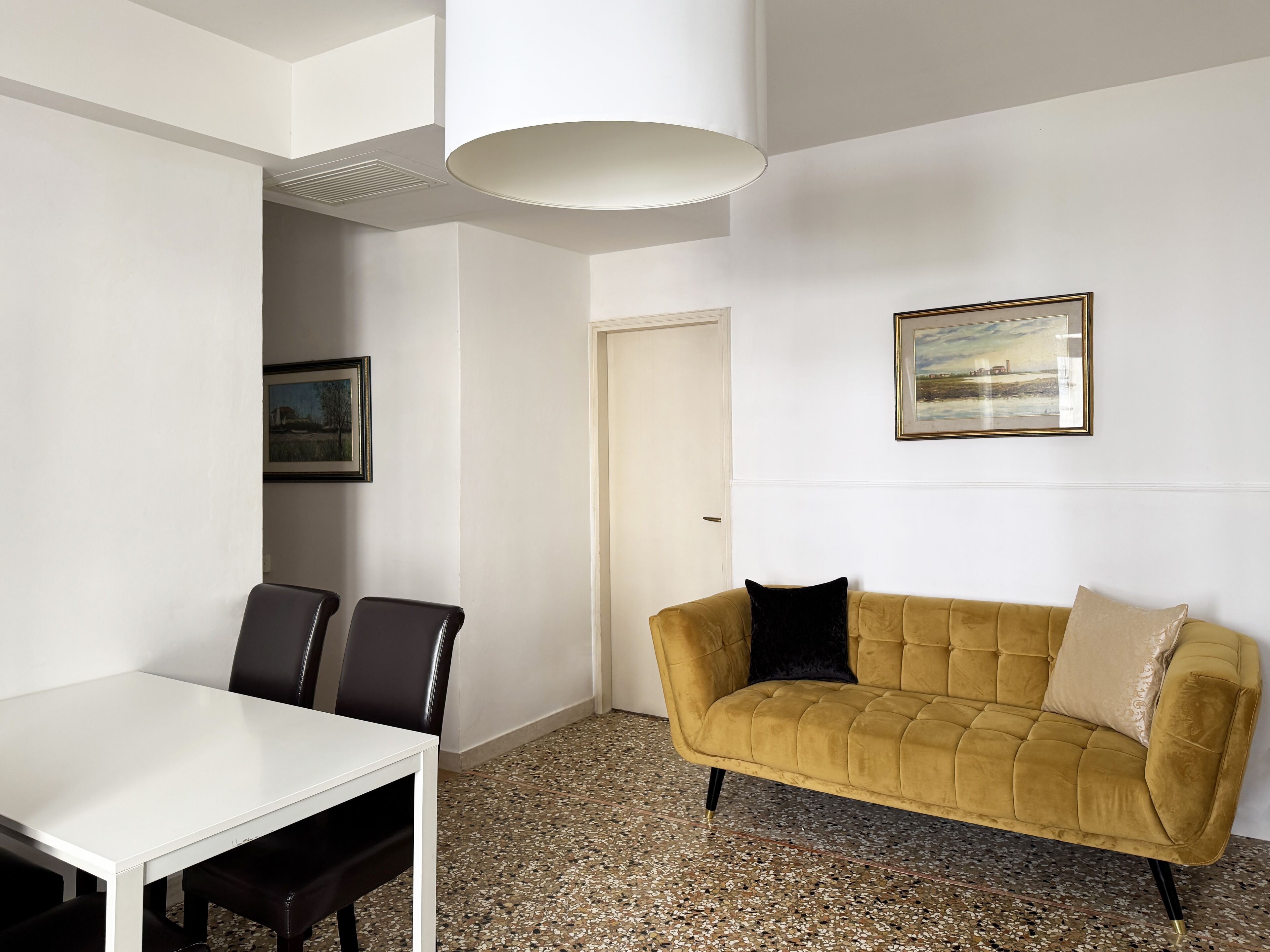 A room with a white table and chairs, a yellow sofa with a black pillow, and a framed picture on the wall.
