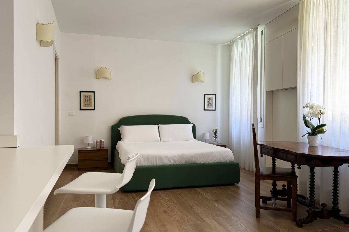 A modern hotel room with a green bed, white walls, and a wooden floor. There is a desk with a chair, a small table with a vase of flowers, and two bedside lamps.
