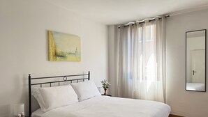 Uma cama bem arrumada com lençóis brancos, uma pequena mesa de cabeceira com abajur, um quadro na parede e um espelho grande.