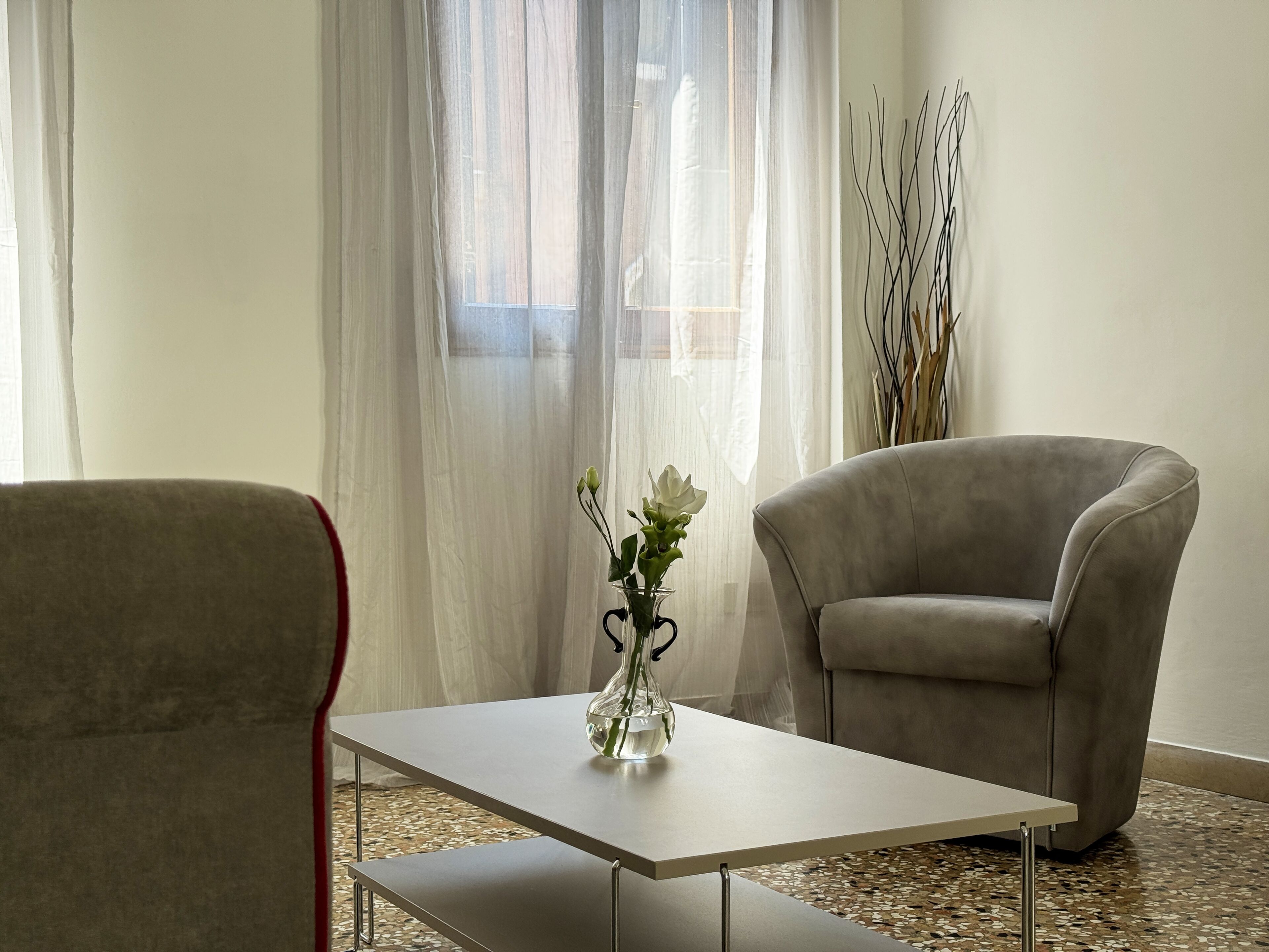 A modern living room with a grey armchair, a small table with a vase of flowers, and sheer curtains.
