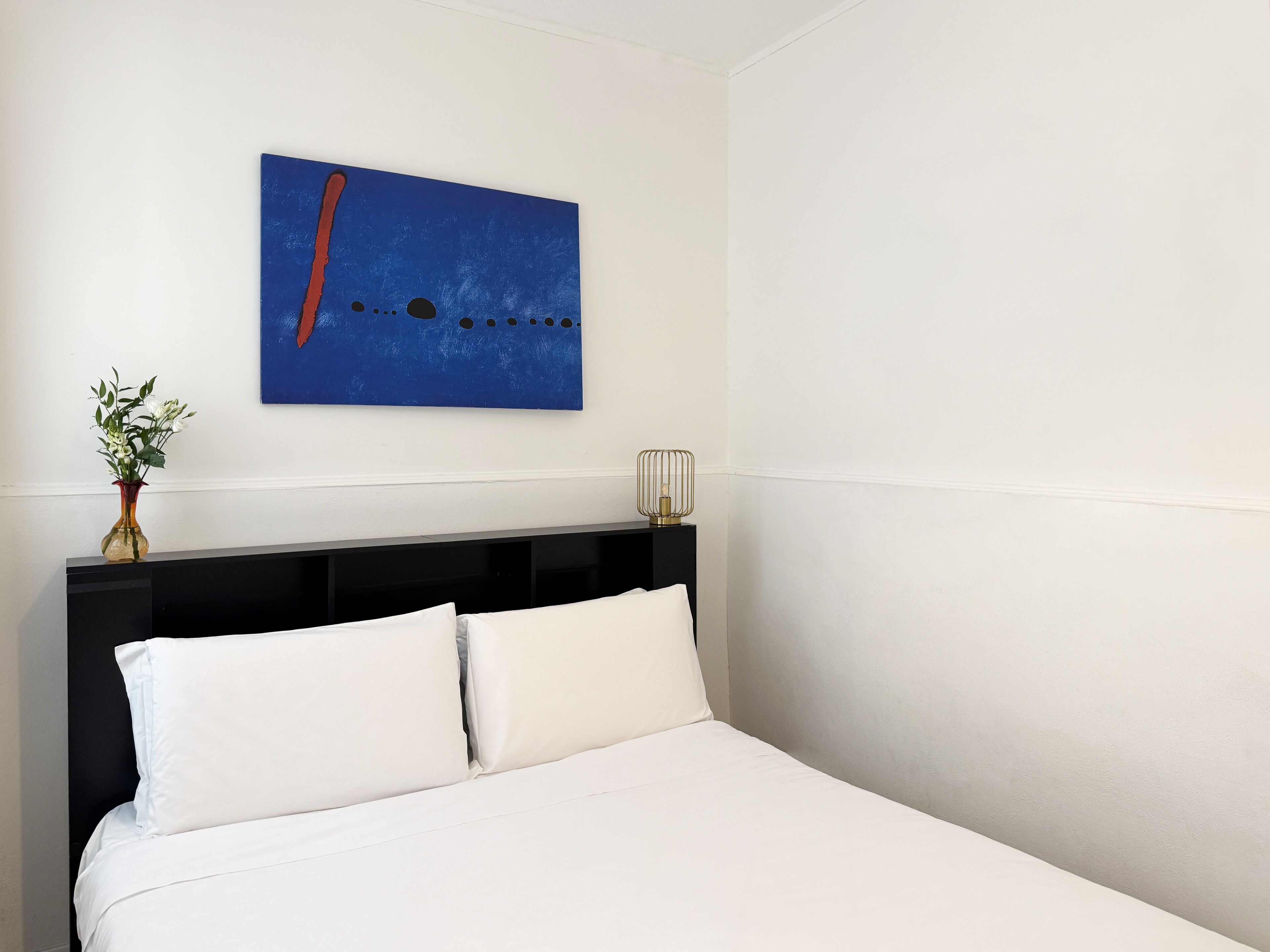 Een net opgemaakt bed met witte lakens, een zwart hoofdbord en een blauw abstract schilderij erboven.