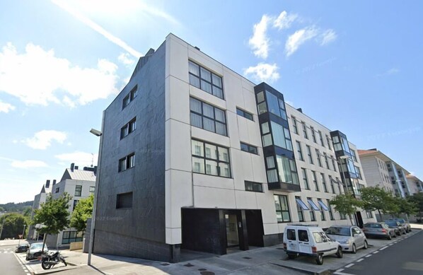 A modern multi-story building with a flat roof, large windows, and a parking area with several cars and a motorcycle.