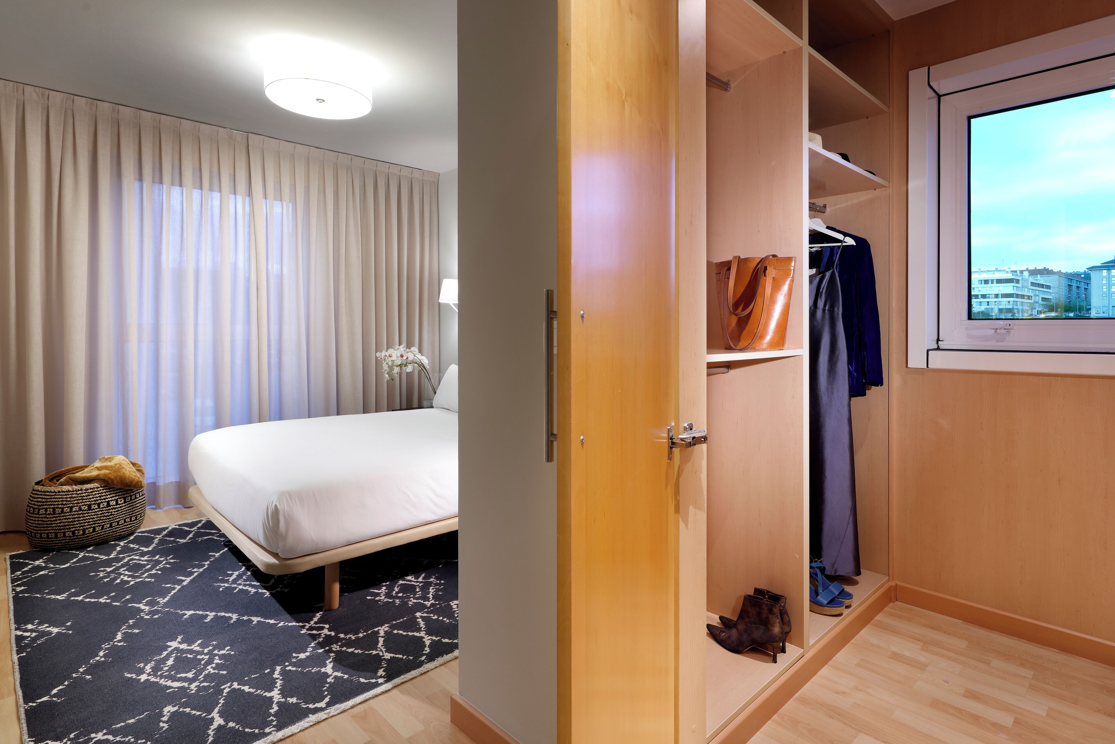 Een hotelkamer met een bed, een kledingkast, een raam met uitzicht over de stad, en een kamerbreed tapijt met een patroon.