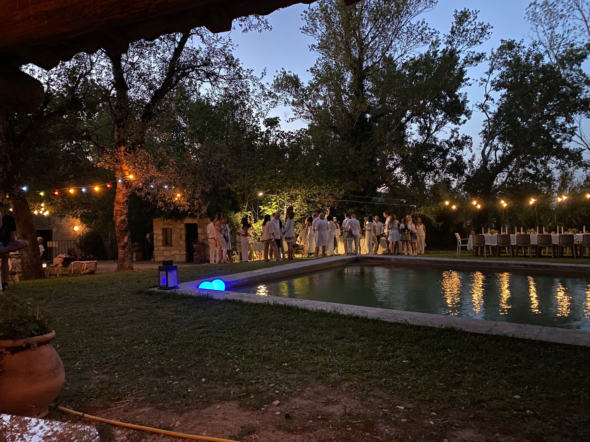 An outdoor event with a pool, string lights, and a gathering of people.