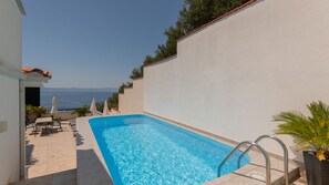 A rectangular swimming pool with a clear blue water, surrounded by a white wall and a tiled deck.