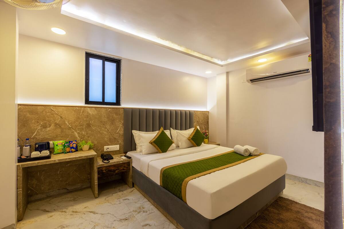 Una habitación de hotel moderna con cabecera de mármol, un escritorio con servicios, una cama grande con una manta verde y un equipo de aire acondicionado.