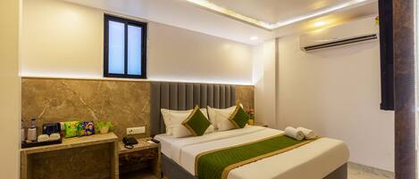 A modern hotel room with a marble headboard, a desk with amenities, a large bed with a green throw, and an air conditioning unit.