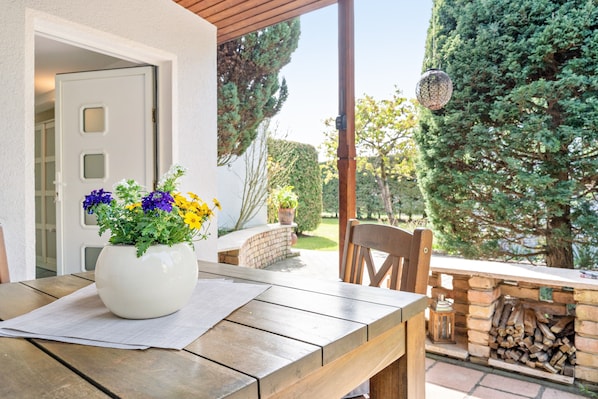 An outdoor patio with a wooden table, a white vase with flowers, a brick fireplace, and a view of a garden.