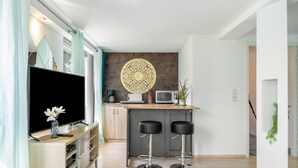 A modern kitchen with a flat-screen TV, two bar stools, a decorative wall hanging, and a small island with a microwave.