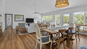 A spacious dining area with a wooden table and chairs, a large woven light fixture, and a view of greenery through large windows. - Shiloh Cottage in Atlantic Beach, NC (Atlantic Beach)