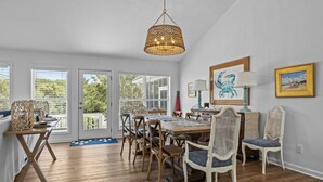 A dining area with a wooden table and chairs, a wicker chandelier, and a view through large windows. - Shiloh Cottage in Atlantic Beach, NC (Atlantic Beach)