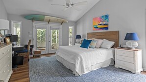 A bedroom with a bed, a desk, a chair, a ceiling fan, a surfboard, and a colorful painting. - Shiloh Cottage in Atlantic Beach, NC (Atlantic Beach)