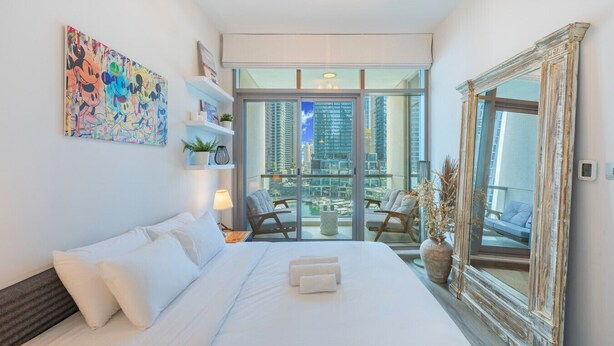 A hotel room with a large bed, a balcony with a view of the city, and a decorative mirror. - Iris Blue - Colourful One Bedroom in The Marina (Dubai Marina)