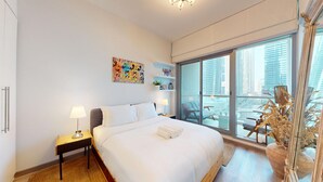 A modern hotel room with a large bed, a bedside table with a lamp, a view of the city through floor-to-ceiling windows, and a decorative plant. - Iris Blue - Colourful One Bedroom in The Marina (Dubai Marina)
