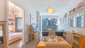 A modern dining area with a wooden table set for four, a city view through large windows, and a cozy seating area with a sofa and a lamp. - Iris Blue - Colourful One Bedroom in The Marina (Dubai Marina)