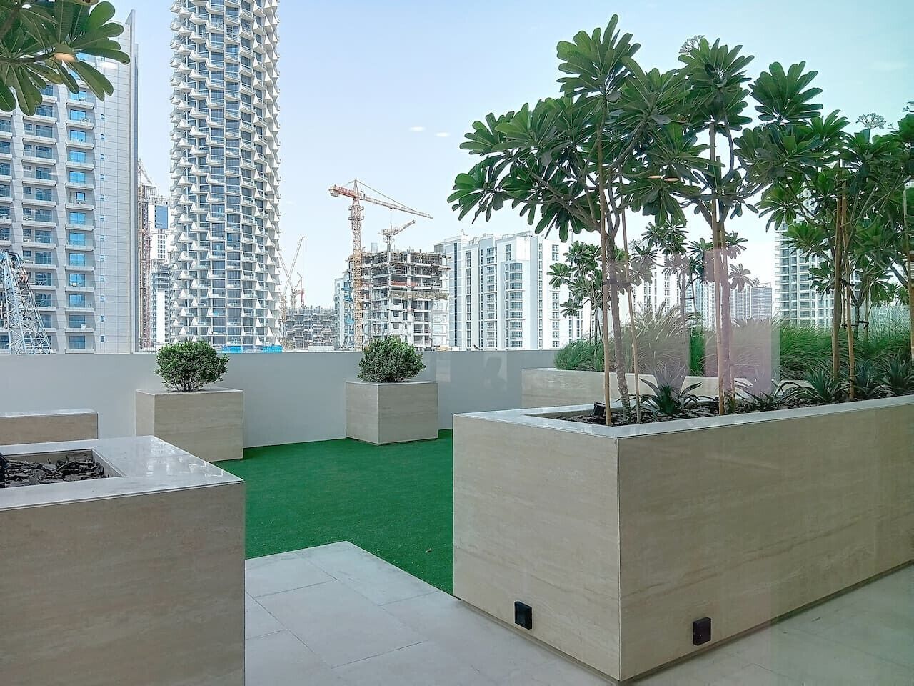 A rooftop garden with green artificial turf, planters with trees and shrubs, and a view of surrounding high-rise buildings and construction cranes.
