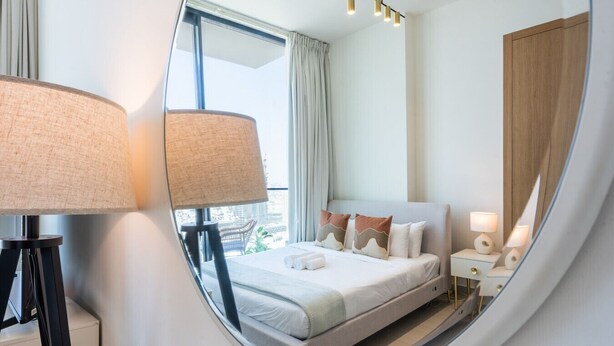 A modern hotel room with a bed, bedside lamps, and a view of the city through large windows.