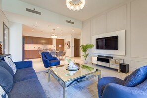 A modern living room with a flat-screen TV, blue sofas, a glass coffee table, and a kitchen in the background. - Vibrant 2 Bedroom l Pool with Burj Khalifa View (Downtown Dubai)