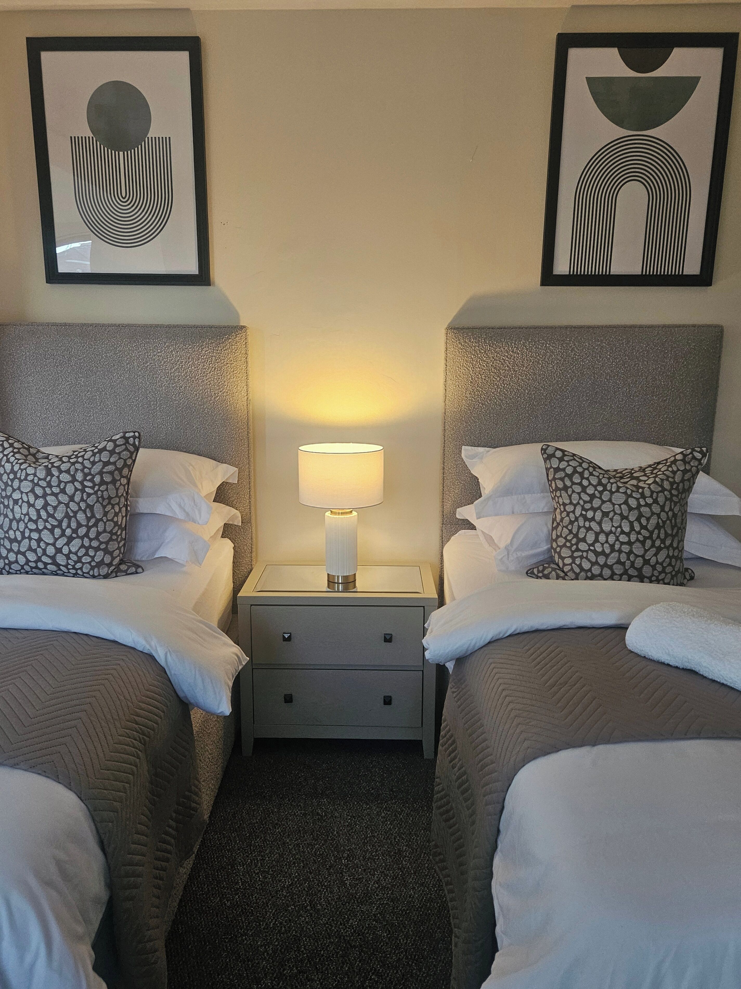 A hotel room with two beds, a nightstand with a lamp, and artwork on the walls.
