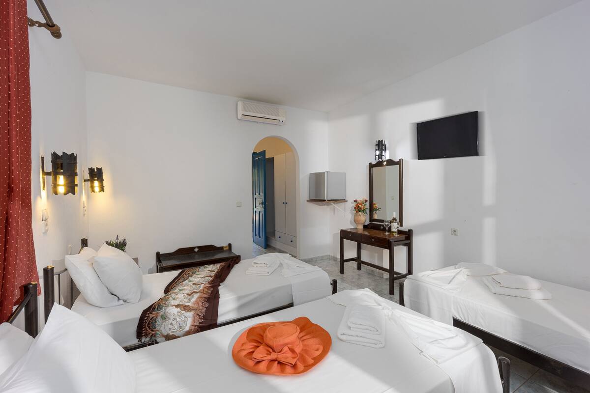 Habitación de hotel con dos camas individuales, un escritorio, una silla, televisión y un baño visible a través de un arco.